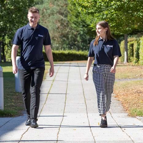 Roke employees wearing navy blue Roke polo shirts walk down a path at the Romsey site. One is male and one is female.
